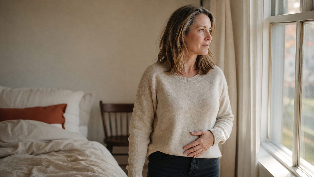 Woman in her late 40s standing by a bedroom window in the morning, one hand resting on her lower stomach