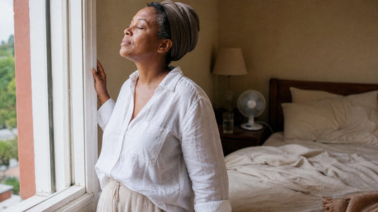 Woman in her late 40s in a white linen sleep shirt breathing fresh air at an open bedroom window at dawn, a fan and glass of water by the rumpled bed