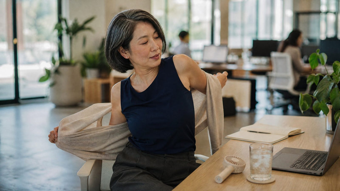 Woman in her early 50s at her office desk slipping off a light cardigan over a sleeveless navy top, with a small fan and iced water beside her laptop
